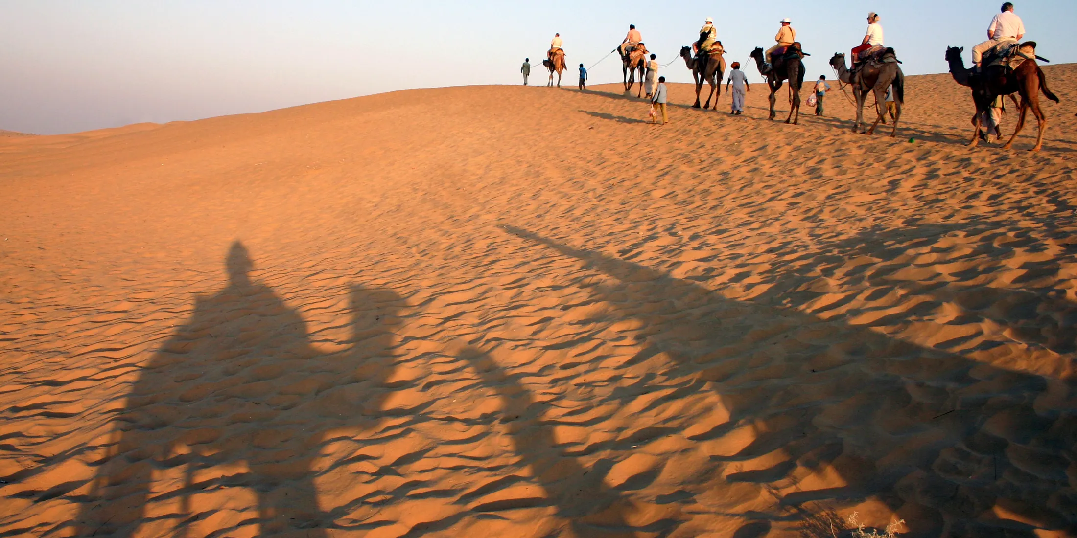 A camel caravan carrying riders up a sunlit dune in the Thar desert